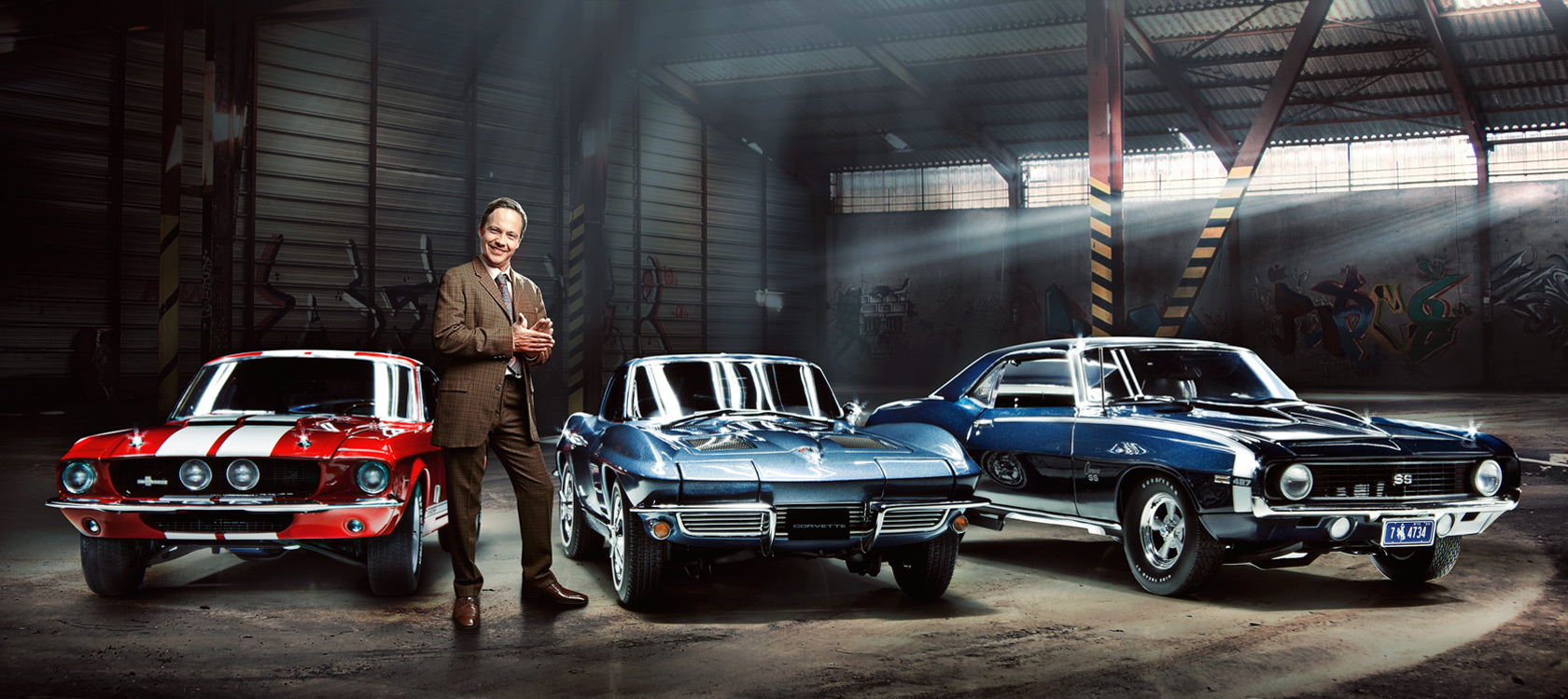 Autoverkäufer im karierten Anzug vor 3 Muscle Cars